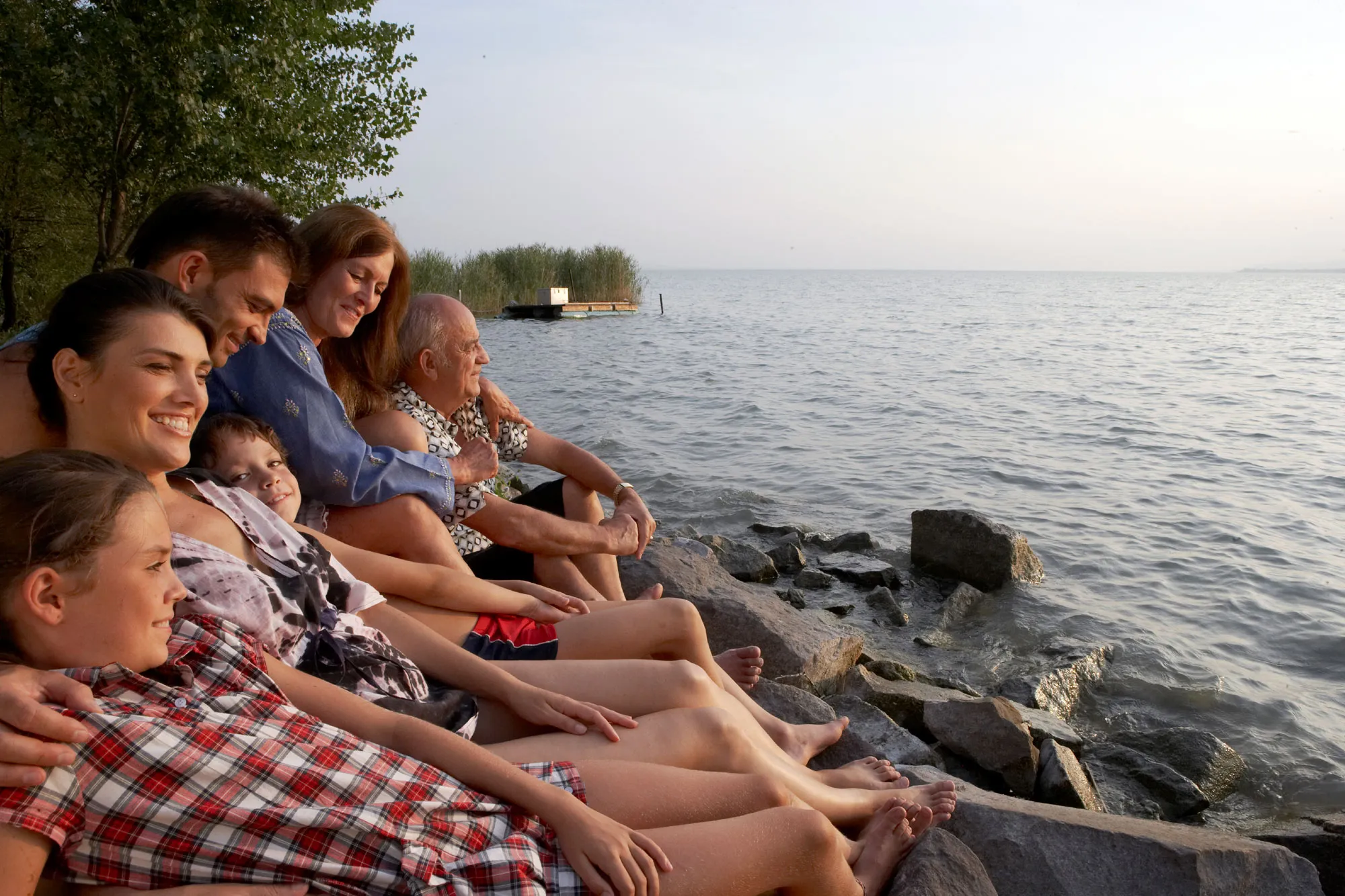 A family of six sits on rocks by a peaceful lake, smiling and embracing warmly under a soft evening sky. Trees and reeds are visible in the background.