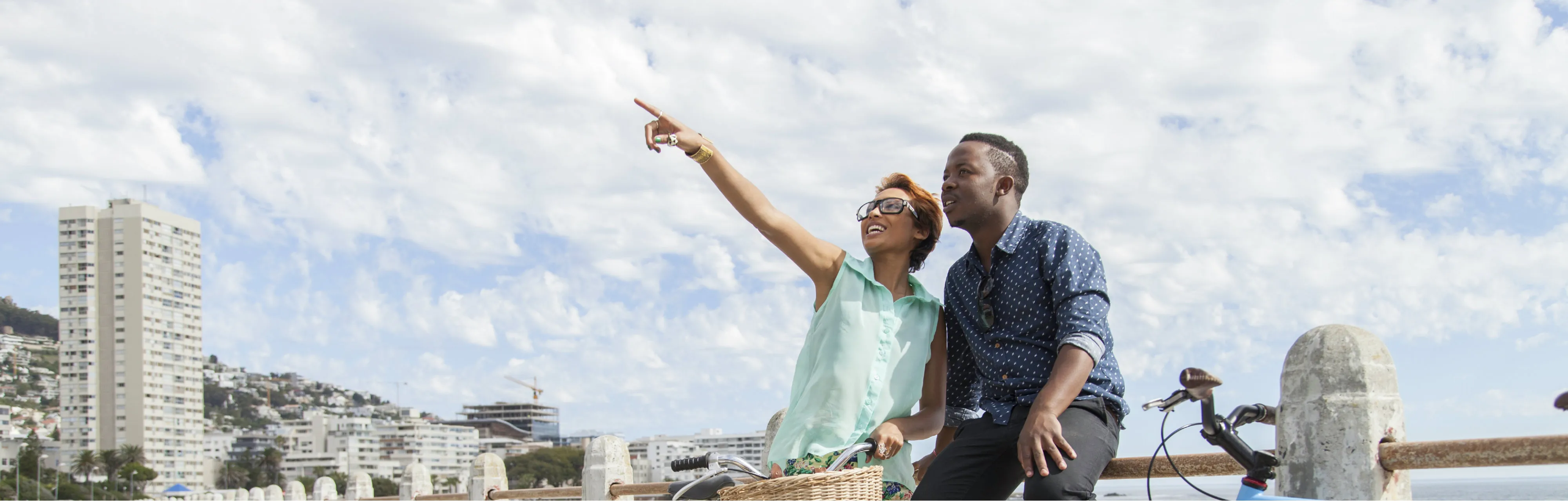 A couple sits on a concrete railing with their bicycles, enjoying a scenic view. The woman points towards the sky, both smiling, under a cloudy blue sky.